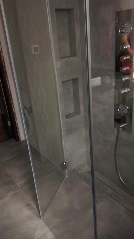 Szklana kabina prysznicowa, z widocznymi elementami montażowymi, na tle szarej ściany z wnękami i panelu prysznicowego z dyszami.