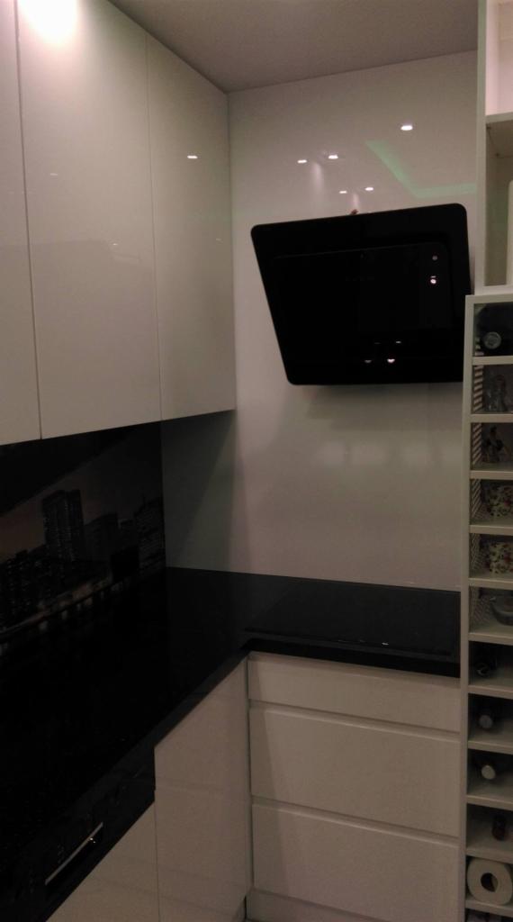 Nowoczesna kuchnia z białymi szafkami na wysoki połysk, czarnym blatem i panelem szklanym z motywem miejskim oraz czarnym okapem.