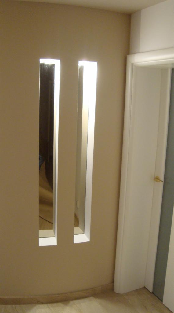 Dwa pionowe, wąskie lustra wpuszczone w beżową ścianę, z białymi ramami, obok białych drzwi z złotą klamką.