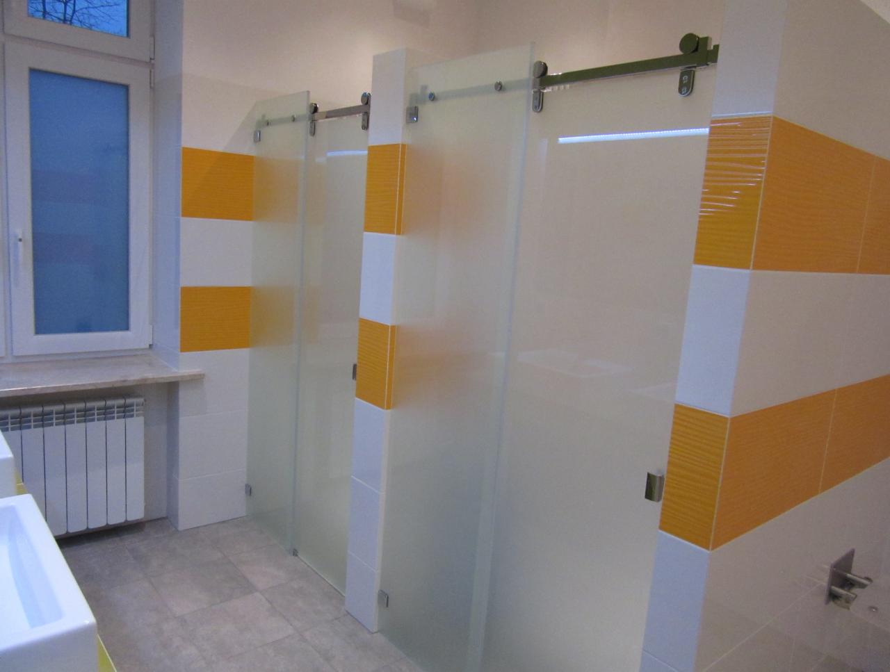 Szklane ścianki działowe w łazience z przesuwnymi drzwiami na szynach, z elementami dekoracyjnymi w kolorze żółtym i białym, widoczne płytki na ścianach i grzejnik pod oknem.