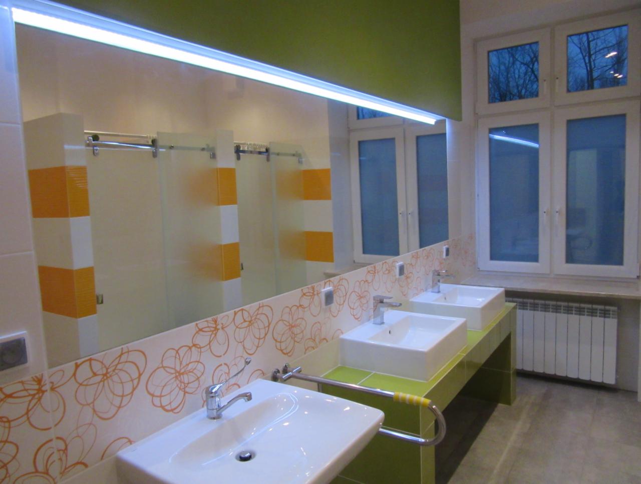 Nowoczesna łazienka z trzema umywalkami, dużym lustrem z oświetleniem LED i kabinami prysznicowymi z pomarańczowymi akcentami, widok na okno zza umywalek.