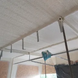 Poddasze z widoczną konstrukcją stalową i świeżo nałożoną, nierówną warstwą białej pianki izolacyjnej, z fragmentami niezabezpieczonych okien w tle.