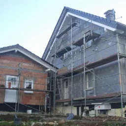 Budowa domu: ściany z cegły i ocieplenie na rusztowaniach, widoczne okna i drzwi, częściowo wykonane pokrycie dachu.