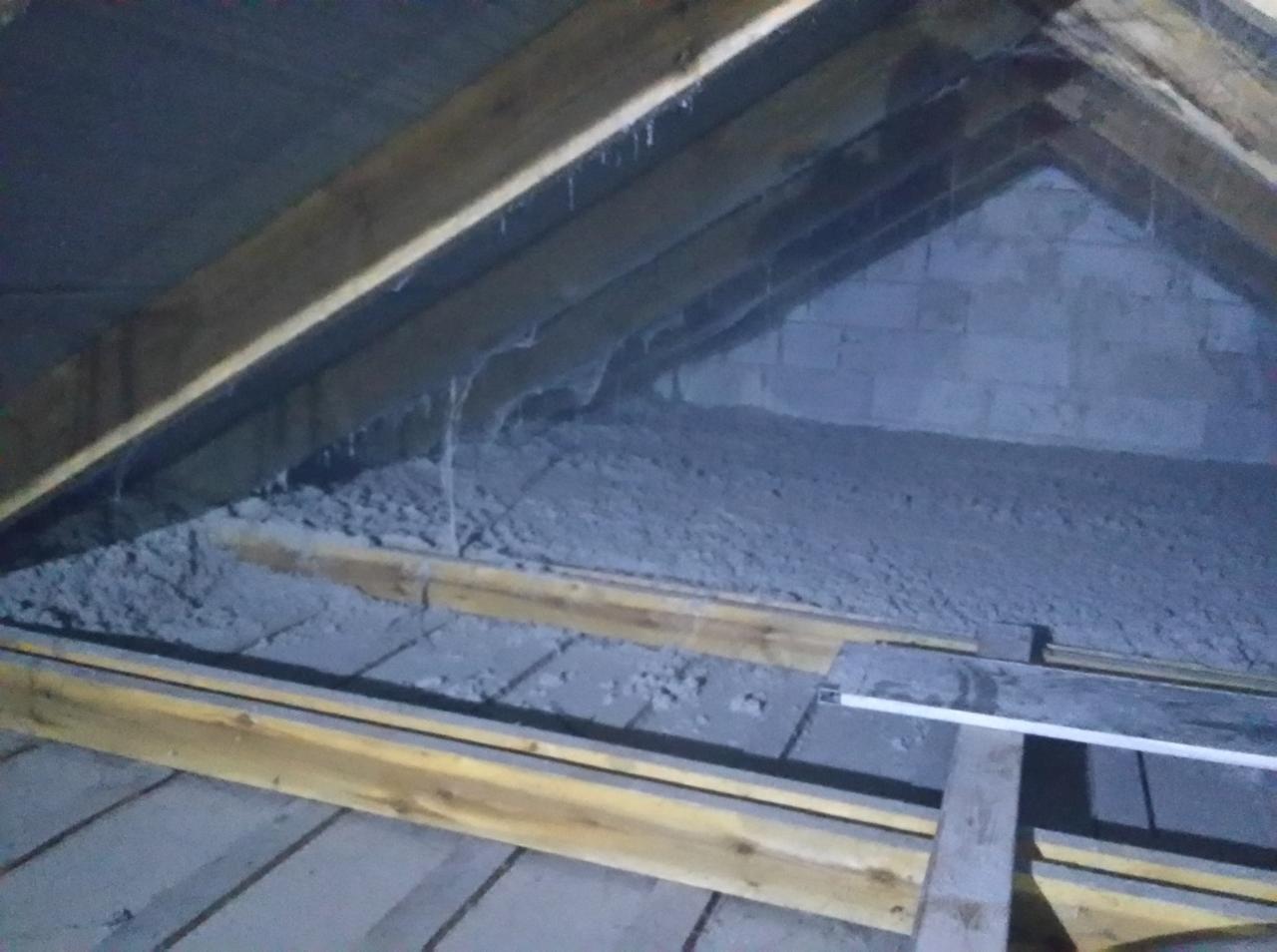 Szara, włóknista warstwa izolacyjna wypełnia przestrzeń poddasza między drewnianymi krokwiami i ścianą z pustaków. Widoczne fragmenty drewnianej konstrukcji dachu i podłogi poddasza.