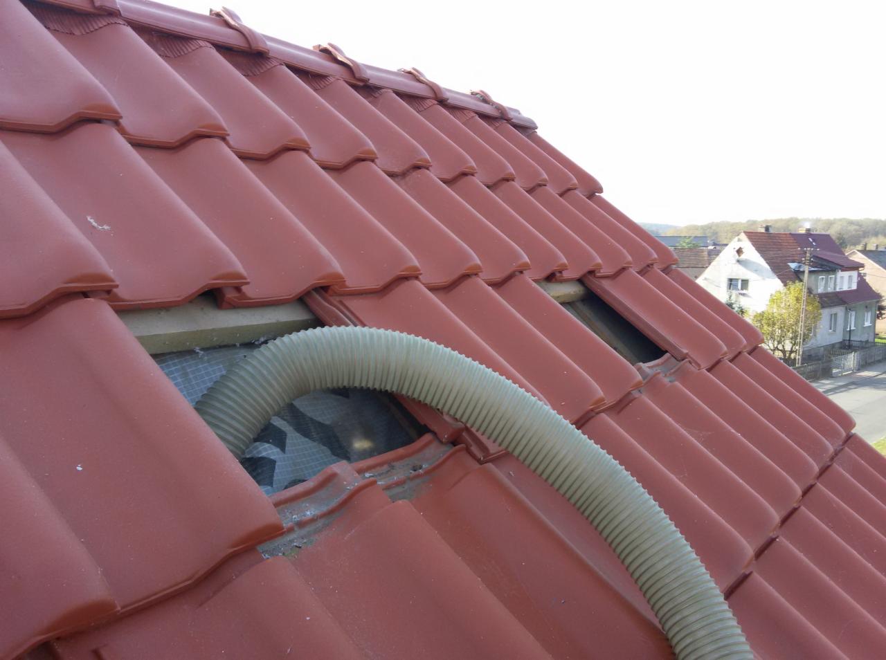 Czerwona dachówka ceramiczna z widoczną przerwą i elastycznym przewodem wentylacyjnym, sugerującym prace związane z wentylacją lub izolacją poddasza.