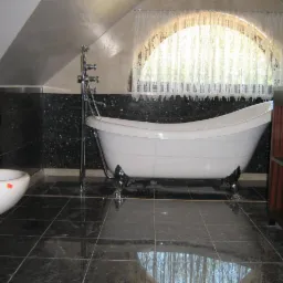 Biała wanna wolnostojąca na chromowanych nóżkach w łazience z czarną, błyszczącą glazurą, widoczne również bidet oraz szafka z ciemnego drewna.