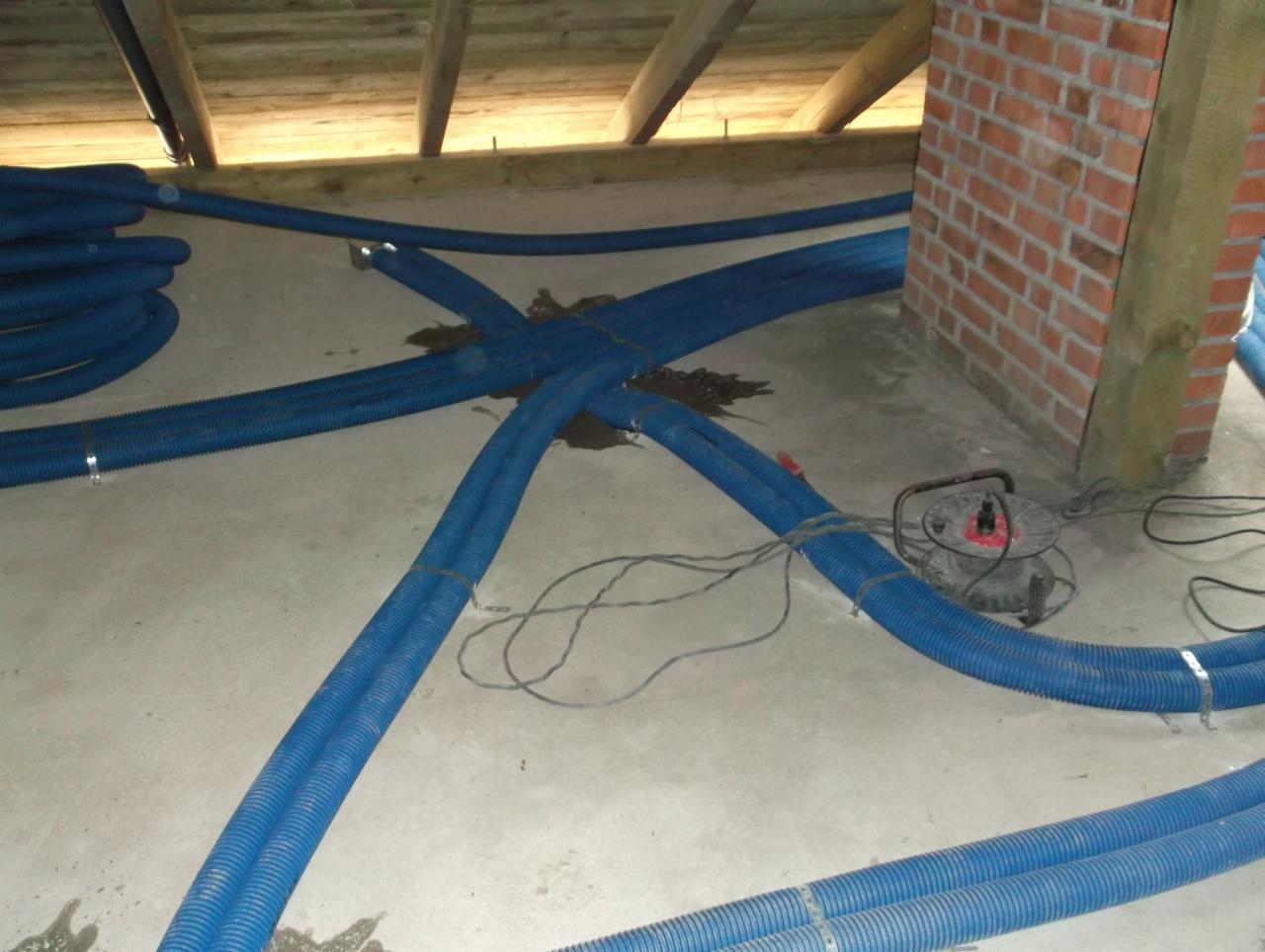 Niebieskie rury karbowane układane na szarej betonowej podłodze poddasza z widoczną ceglaną ścianą komina i zwisającymi belkami stropu, prawdopodobnie w trakcie montażu ogrzewania podłogowego.