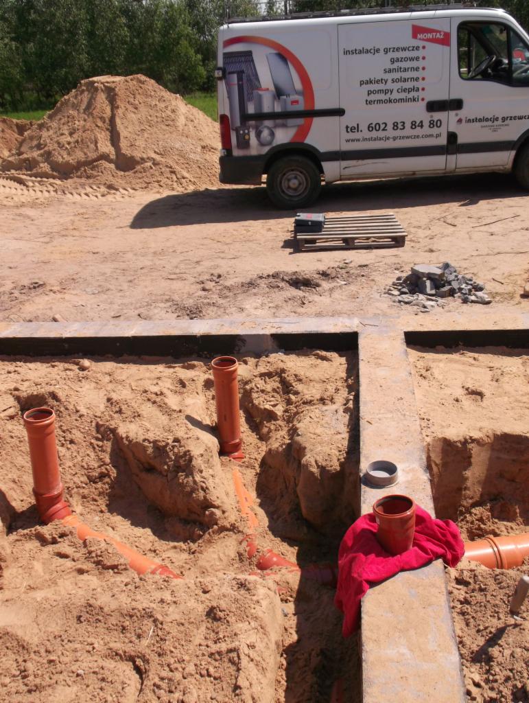 Wykop z pomarańczowymi rurami kanalizacyjnymi w ziemi, obok betonowego fundamentu z czerwoną szmatką i rurą, w tle samochód dostawczy z reklamą instalacji grzewczych.
