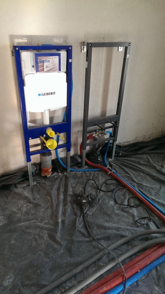 Stelaże podtynkowe WC Geberit Sigma w trakcie montażu, widoczne rury doprowadzające wodę i odpływ, zabezpieczone folią podłoże.