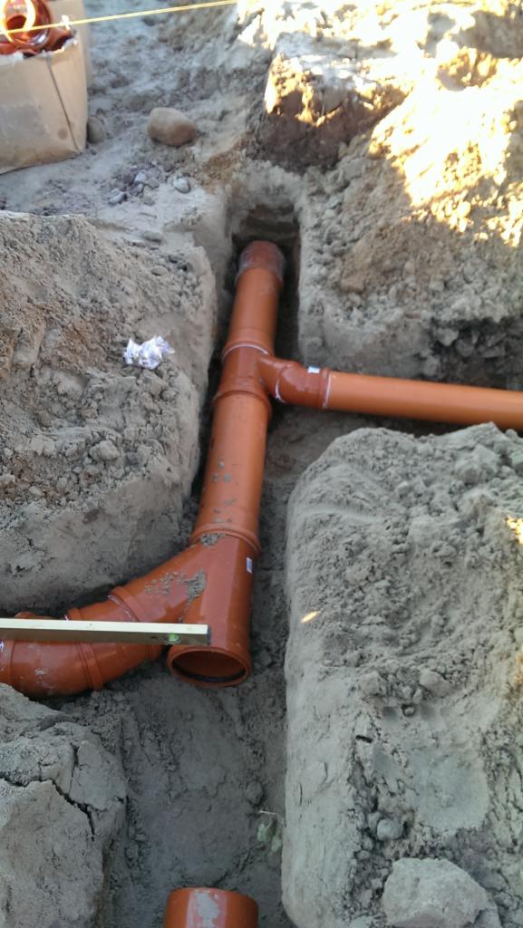 Pomarańczowe rury kanalizacyjne ułożone w wykopie ziemnym, widoczne połączenia i poziomica na jednej z rur.