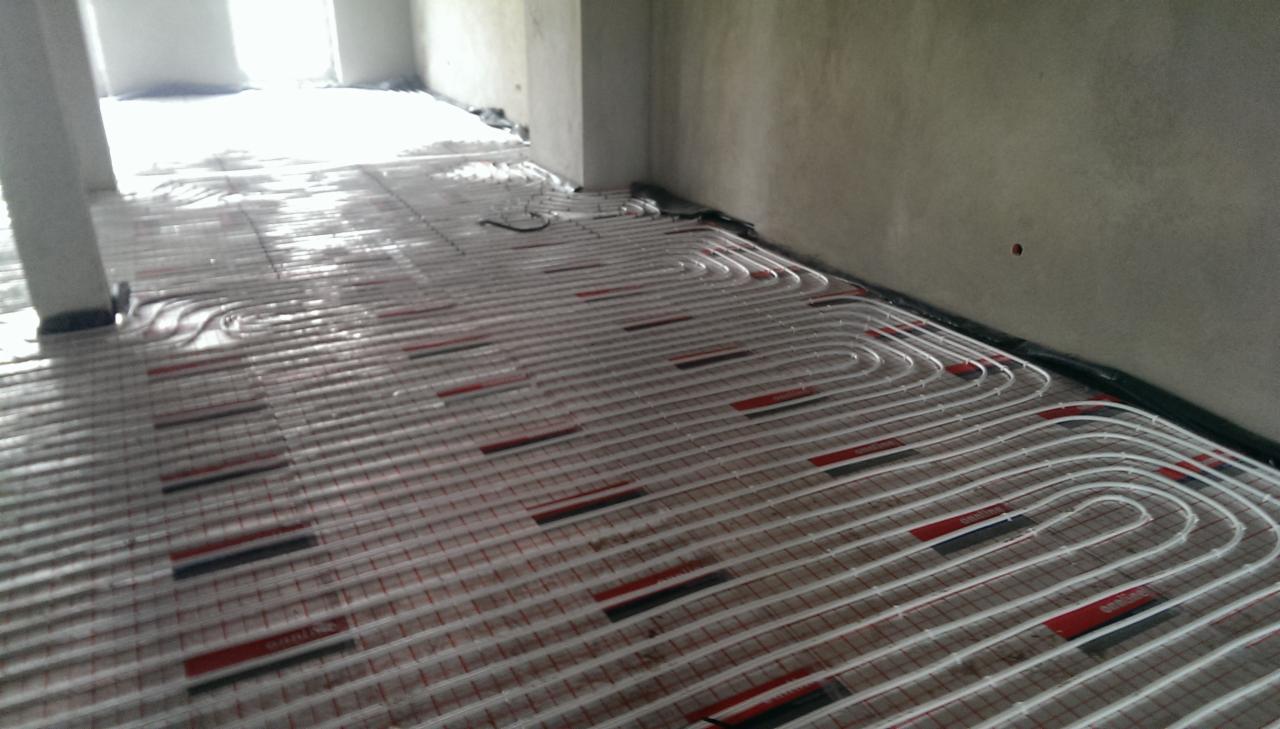Białe rurki ogrzewania podłogowego ułożone równolegle na podłodze z siatką, widoczne czerwone i czarne elementy mocujące, surowy stan ścian i podłoża.
