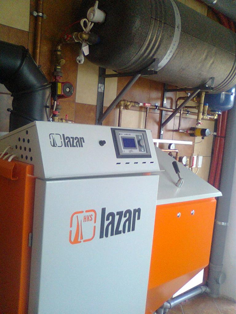 Kocioł grzewczy Lazar z elektronicznym panelem sterowania, obok zbiornik akumulacyjny w stalowej obudowie i instalacja rurowa z zaworami.