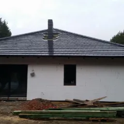 Dom w trakcie budowy w Kotlinie, z widocznym nowym dachem z ciemnej dachówki i kominem, ściany zewnętrzne z białego styropianu, teren wokół domu niezagospodarowany.