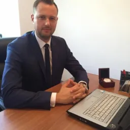 Mężczyzna w garniturze siedzi przy biurku z laptopem, zegarkiem w pudełku i przyborami do pisania, sugerując pracę biurową związaną z finansami.