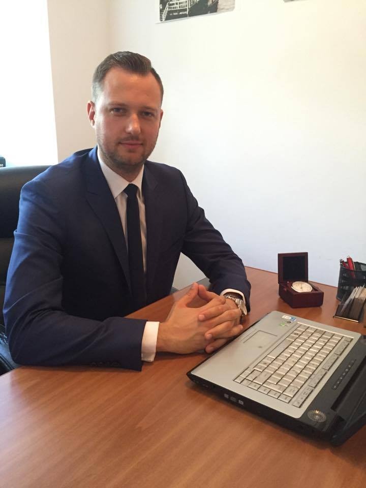 Mężczyzna w garniturze siedzi przy biurku z laptopem, zegarkiem w pudełku i przyborami do pisania, sugerując pracę biurową związaną z finansami.