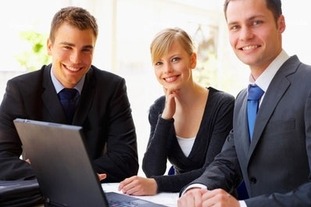 Trzy osoby uśmiechają się do kamery, siedząc przy stole z laptopem. Mężczyzna po lewej ma na sobie ciemny krawat w niebieskie wzory.