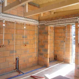 Wnętrze w stanie surowym z widoczną instalacją elektryczną w peszlach i rurach wodno-kanalizacyjnych, ściany z cegły dziurawki, drewniane belki stropowe.