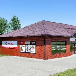 Budynek firmy Vetrex z brązową dachówką, prezentujący okna, drzwi, schody, bramy, rolety i parapety w oknach wystawowych oraz plakaty reklamowe okien w oknach