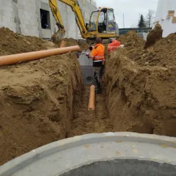 Montaż betonowej studzienki kanalizacyjnej w wykopie; pracownik w odblaskowej kurtce łączy elementy, w tle koparka i budynek w trakcie budowy.