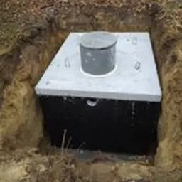 Betonowy zbiornik szamba, częściowo zagłębiony w ziemi, z widocznym włazem i czterema uchwytami montażowymi na pokrywie, otoczony wykopem w ziemi, w tle las.