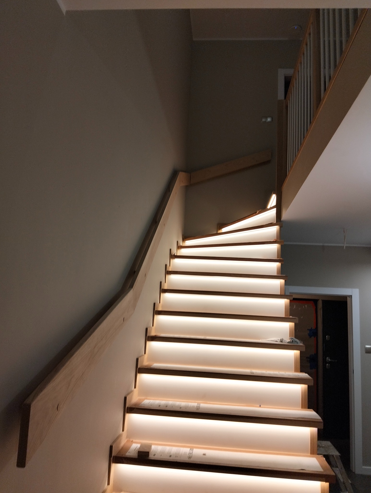 Nowoczesne schody z podświetleniem LED, drewniane stopnie i jasna ściana. Widok z dołu, ukazujący zakrzywiony bieg schodów i minimalistyczny design.