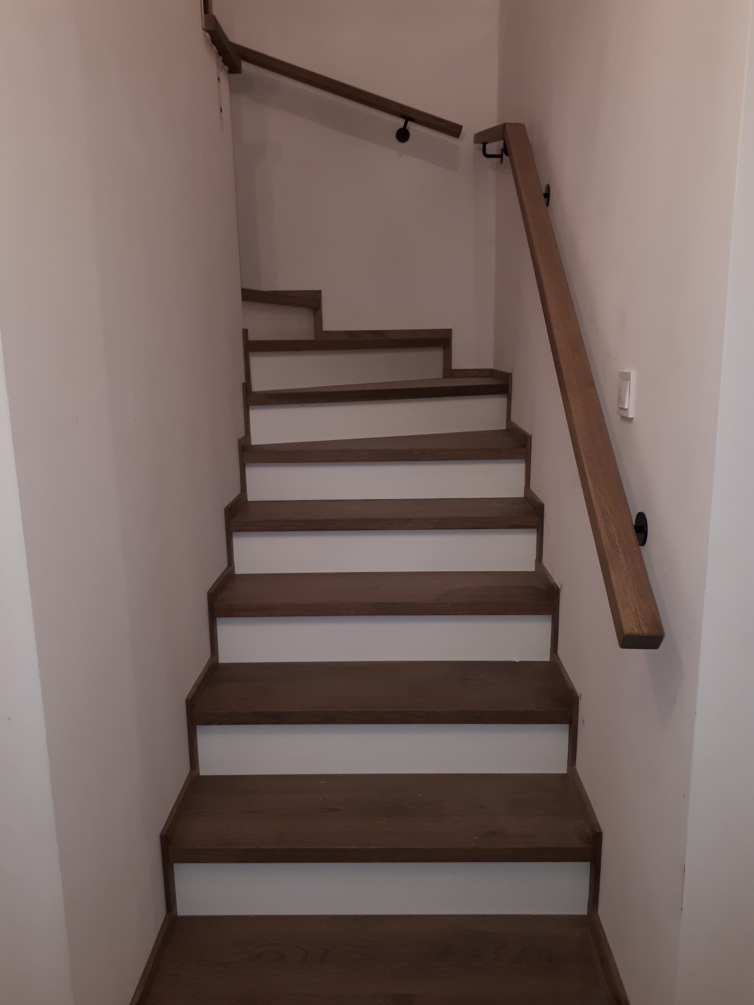 Drewniane schody z brązowymi stopniami i białą podstopnicą, z drewnianą poręczą zamocowaną do ściany. Widok z dołu na górę.