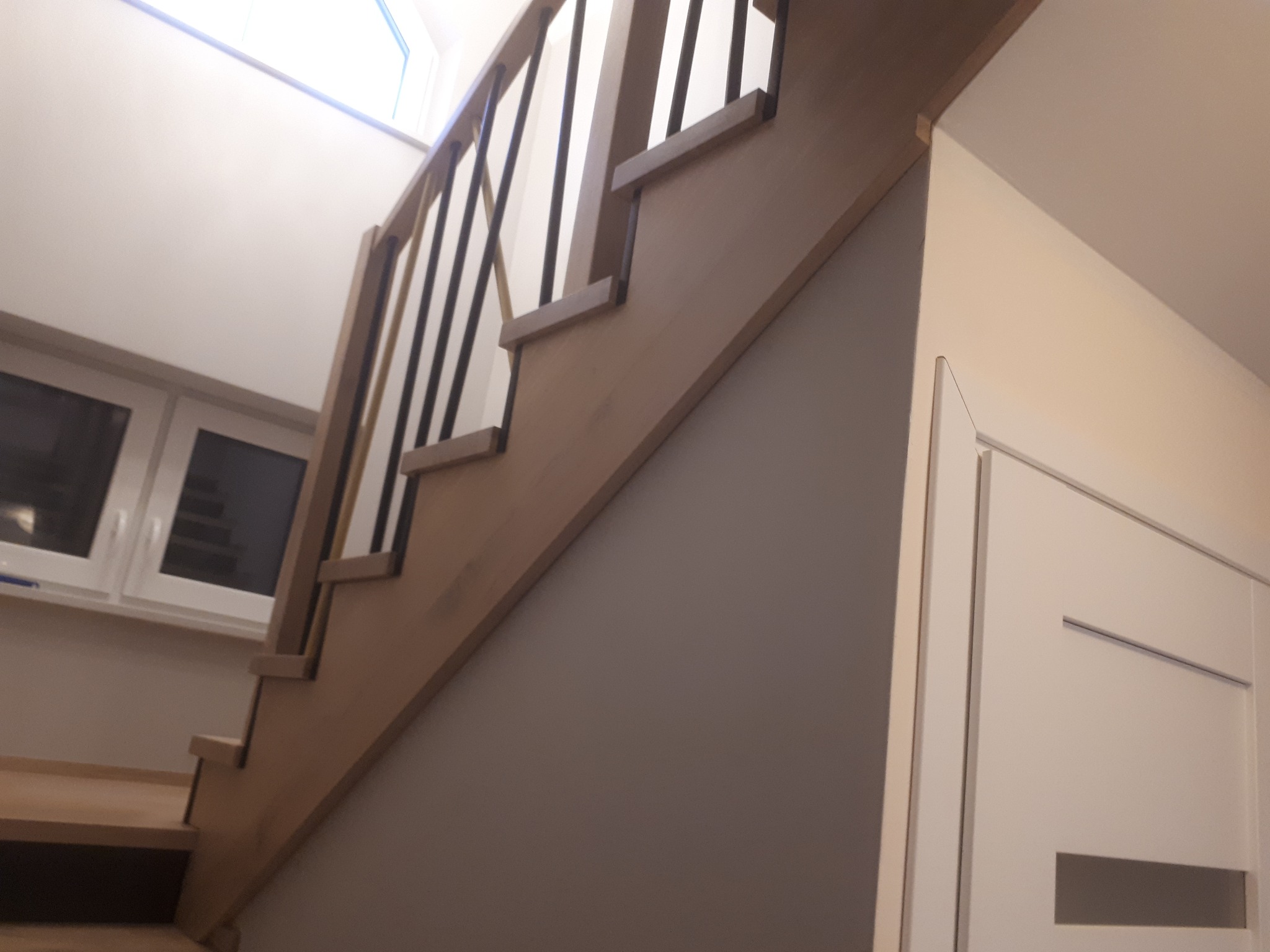 Jasne schody z ciemnymi szczeblami balustrady, widoczne od dołu, z fragmentem białych drzwi i oknem w tle. Ujęcie z niskiej perspektywy.