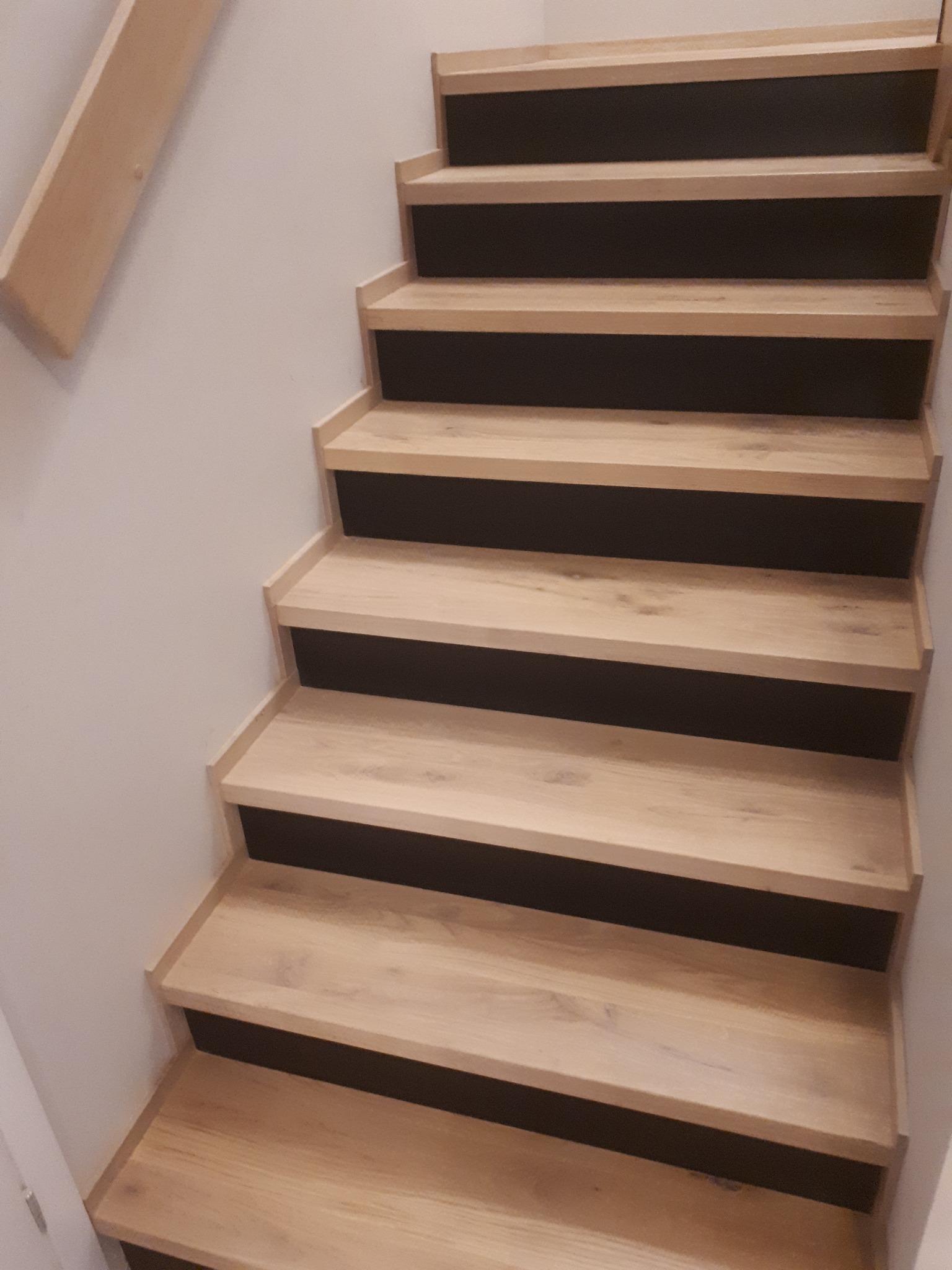 Jasne, drewniane schody z ciemnymi podstopnicami, widok z dołu, ukazujący detale wykończenia i strukturę drewna. Minimalistyczny design.