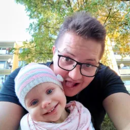 Selfie ojca z uśmiechniętą córeczką w czapce i szaliku na tle drzewa i budynku.