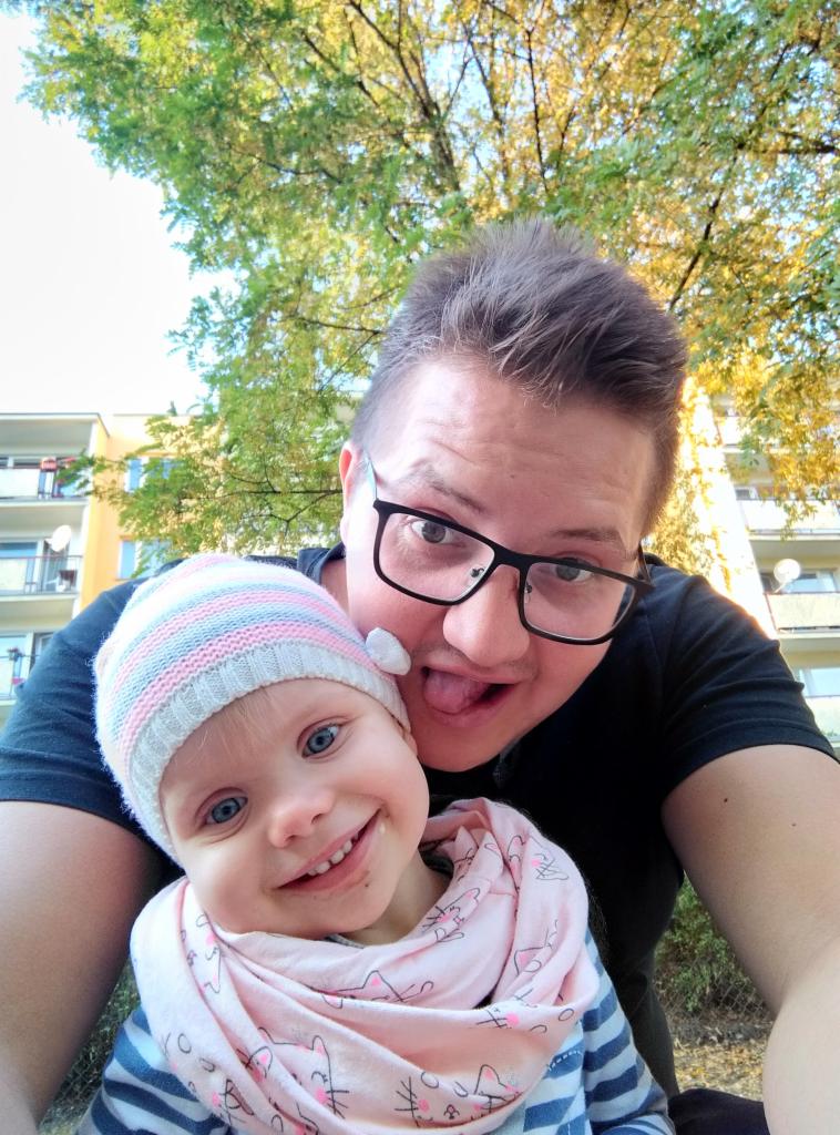 Selfie ojca z uśmiechniętą córeczką w czapce i szaliku na tle drzewa i budynku.