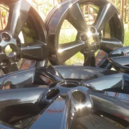 Błyszczące, czarne felgi aluminiowe po renowacji, ułożone na drewnianej powierzchni w plenerze, z widocznymi refleksami słonecznymi na powierzchni.