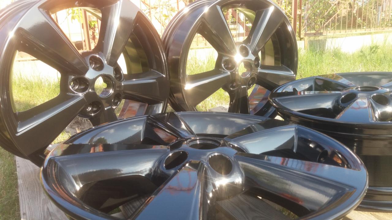 Błyszczące, czarne felgi aluminiowe po renowacji, ułożone na drewnianej powierzchni w plenerze, z widocznymi refleksami słonecznymi na powierzchni.