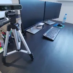 Stanowisko pracy z dwoma monitorami Dell, klawiaturą Logitech, myszką, kamerą internetową na statywie, butelką wody i organizerem na biurko.