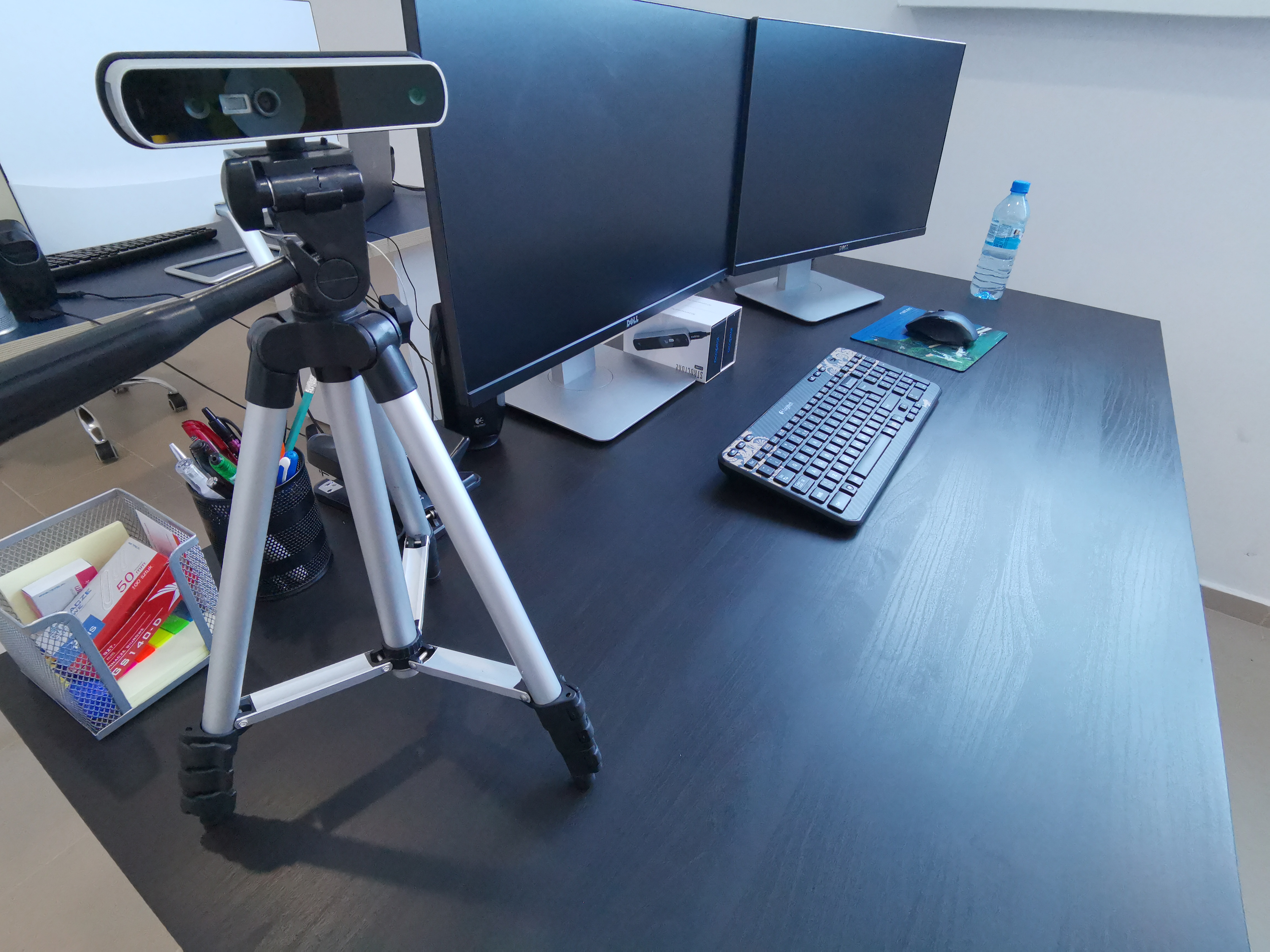 Stanowisko pracy z dwoma monitorami Dell, klawiaturą Logitech, myszką, kamerą internetową na statywie, butelką wody i organizerem na biurko.