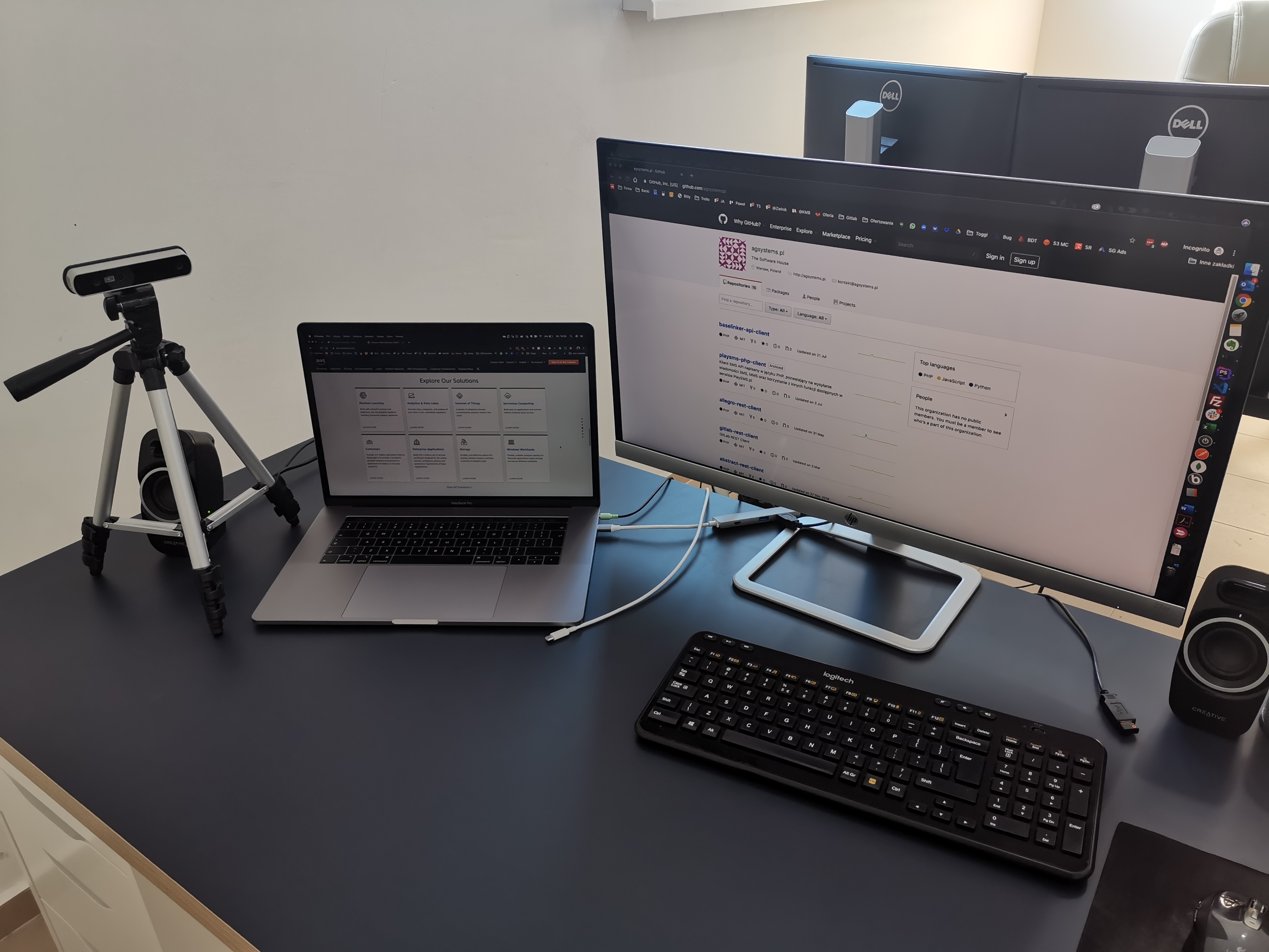 Stanowisko pracy z laptopem z otwartą stroną AWS, monitorem wyświetlającym kod z GitHub, kamerą internetową na statywie, klawiaturą Logitech i głośnikiem Creative.