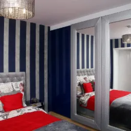 Sypialnia z łóżkiem z szarym wezgłowiem, czerwonymi poduszkami i narzutą, szafa przesuwna z lustrami w srebrnych ramach, ściany w granatowo-srebrne pasy, okrągły żyrandol.