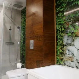 Nowoczesna łazienka z drewnianą szafką na wymiar, wiszącą toaletą i wanną, akcentem zielonej fototapety z motywem kamienia i bluszczu.