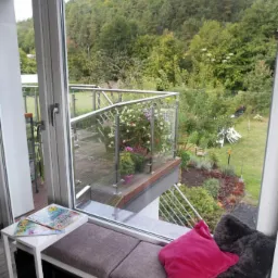 Okno z siedziskiem i widokiem na balkon z kwiatami i zielony ogród w tle.