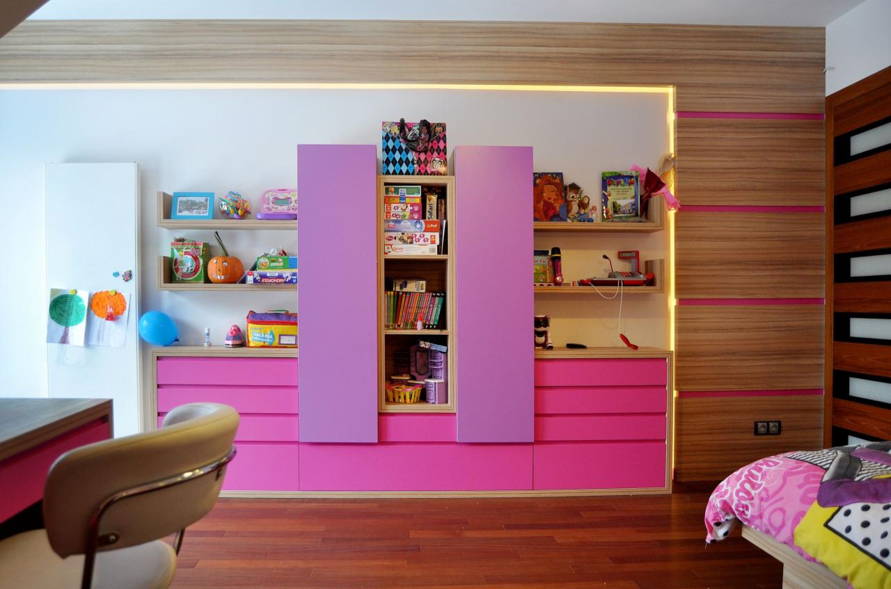 Pokój dziecięcy z zabudową meblową w odcieniach różu i fioletu, zawierający regały z zabawkami, biurko z krzesłem i łóżko z kolorową pościelą, oświetlenie LED podkreśla drewniane wykończenia.
