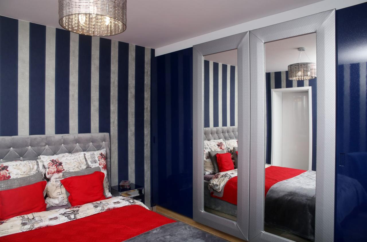 Sypialnia z łóżkiem z szarym wezgłowiem, czerwonymi poduszkami i narzutą, szafa przesuwna z lustrami w srebrnych ramach, ściany w granatowo-srebrne pasy, okrągły żyrandol.
