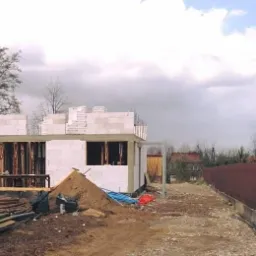 Wznoszenie ścian parterowego domu z betonu komórkowego, widoczne drewniane stemple podtrzymujące strop, na pierwszym planie sterta desek i piasku, w tle ogrodzenie i drzewa.