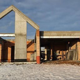 RYŻ-BUD - Surowy stan domu z betonu komórkowego w zimowej scenerii. Widoczne ściany, słupy i nadproża. Na pierwszym planie śnieg, w tle zalesione wzgórze i pochmurne niebo.