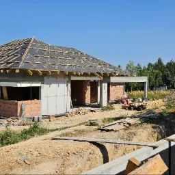 Budowa domu jednorodzinnego w trakcie realizacji: wzniesione mury z pustaków, przygotowane więźby dachowe, widoczne fundamenty i tymczasowa toaleta na placu budowy.