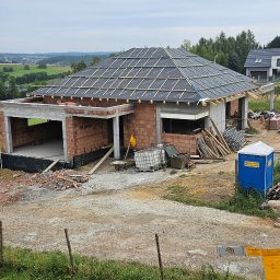 RYŻ-BUD - Budowa domu jednorodzinnego w Krakowie, widok z góry na częściowo ukończony budynek z czerwonej cegły i dachem pokrytym membraną. W tle zielony krajobraz.
