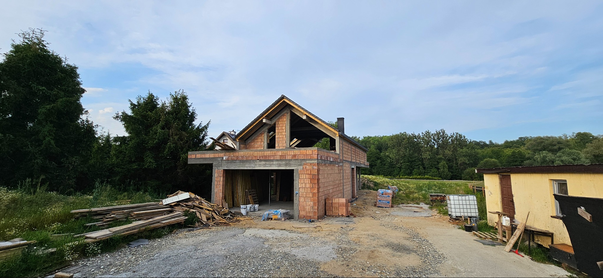 Budowa domu jednorodzinnego w stanie surowym otwartym z czerwonej cegły. Widoczne drewniane elementy konstrukcji dachu i otoczenie z zielenią.