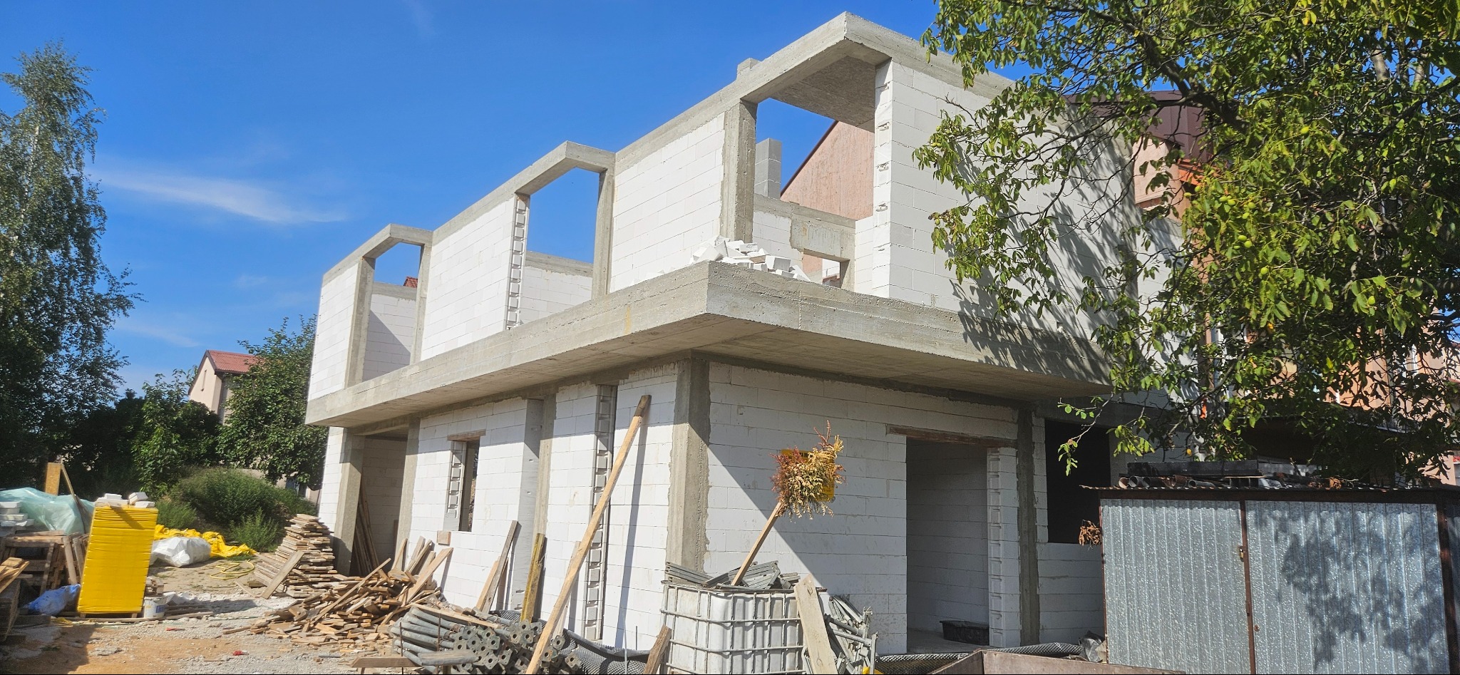 Surowy stan dwukondygnacyjnego domu z betonu komórkowego, widok z zewnątrz, z widocznymi elementami konstrukcyjnymi stropu i ścian, materiały budowlane wokół.