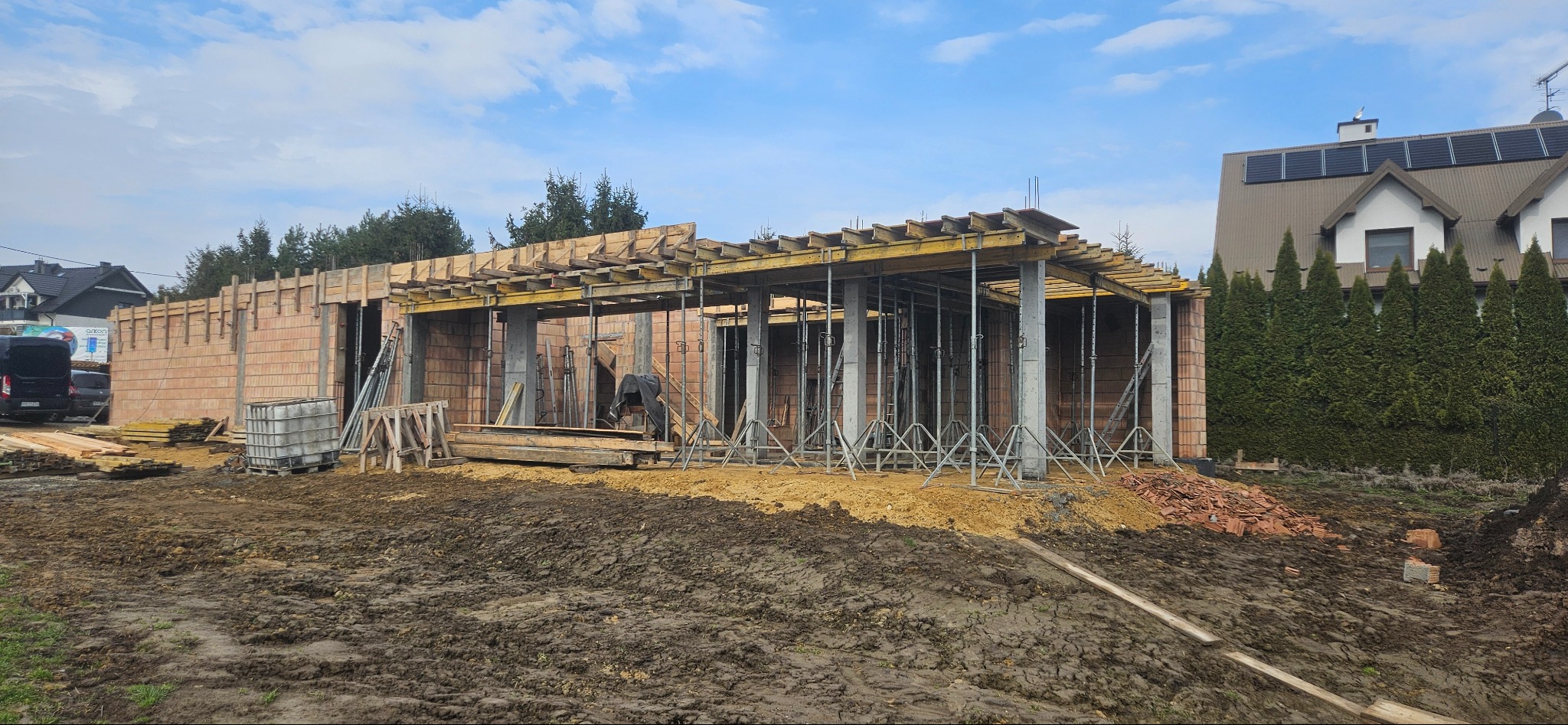 Stan surowy otwarty domu jednorodzinnego w trakcie budowy: ściany z pustaków, betonowe słupy i drewniane szalunki stropu. W tle dom z panelami solarnymi.