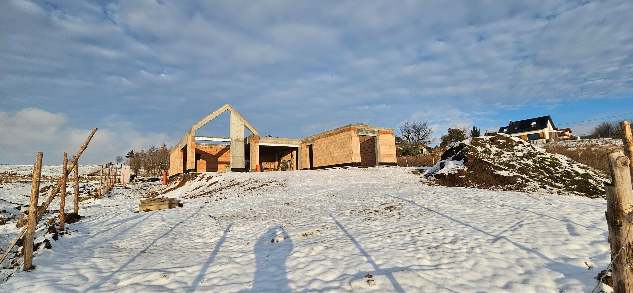 W zimowej scenerii, niedokończony dom z czerwonej cegły z betonowym szczytem, na wzgórzu. W tle widoczny inny dom i błękitne niebo z chmurami.