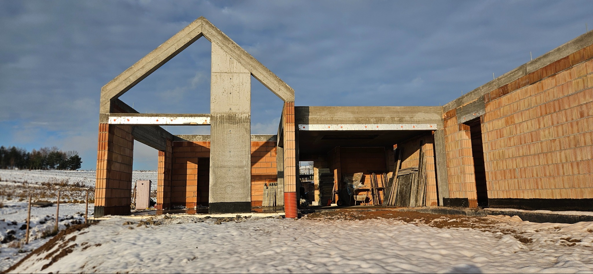 Surowy stan domu z betonu komórkowego w zimowej scenerii. Widoczne ściany, słupy i nadproża. Na pierwszym planie śnieg, w tle zalesione wzgórze i pochmurne niebo.
