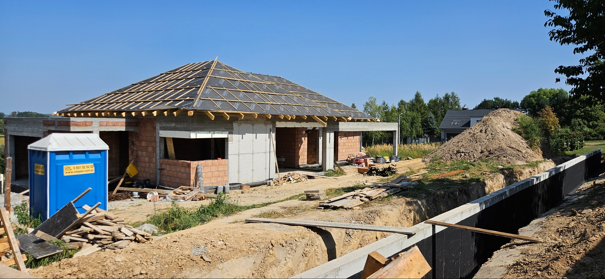 Budowa domu jednorodzinnego w trakcie realizacji: wzniesione mury z pustaków, przygotowane więźby dachowe, widoczne fundamenty i tymczasowa toaleta na placu budowy.